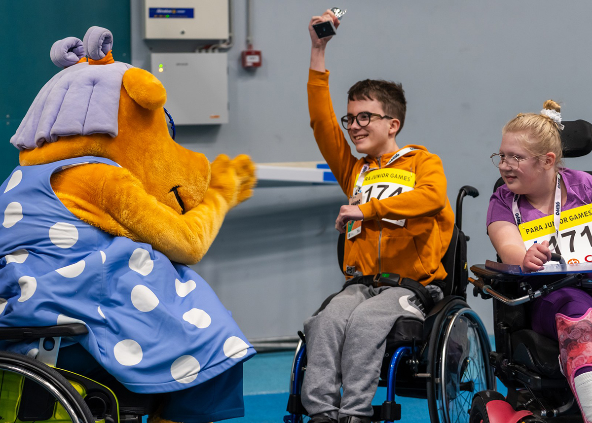 Hippo-hahmo kannustaa pyörätuolia käyttäviä nuoria, jotka tuulettavat pienen palkintopokaalin kanssa.